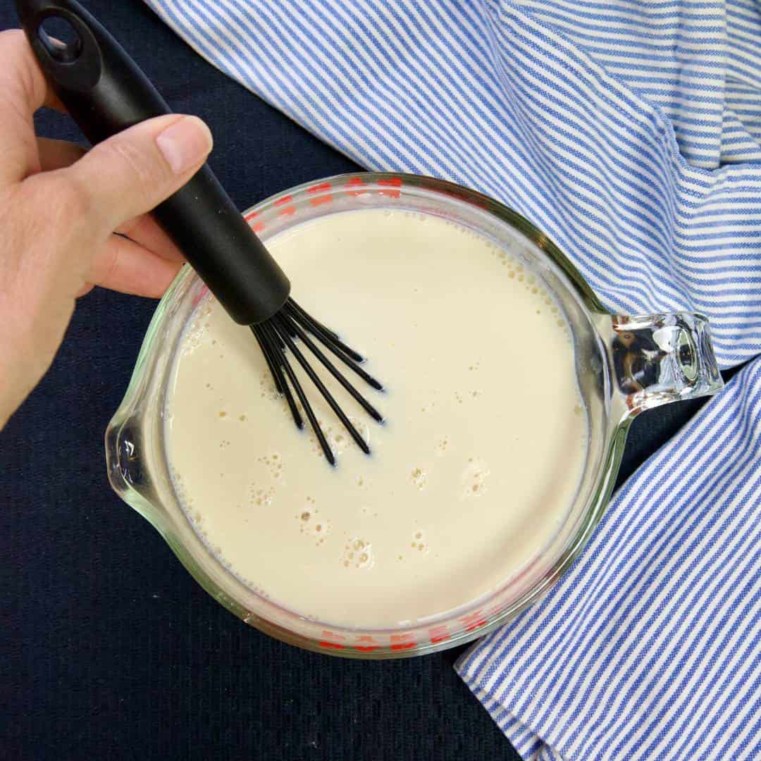 hand holding a whisk in the measuring cup with soy milk striped blue kitchen towel draped next to cup
