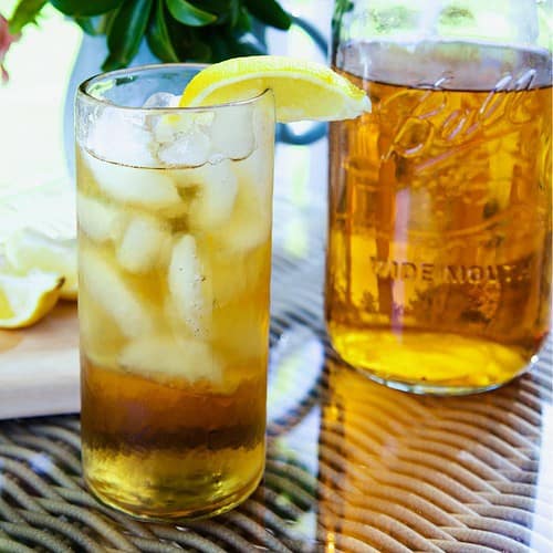 a tall glass of sun tea with ice and a lemon wedge flowers and large mason jar with tea in background