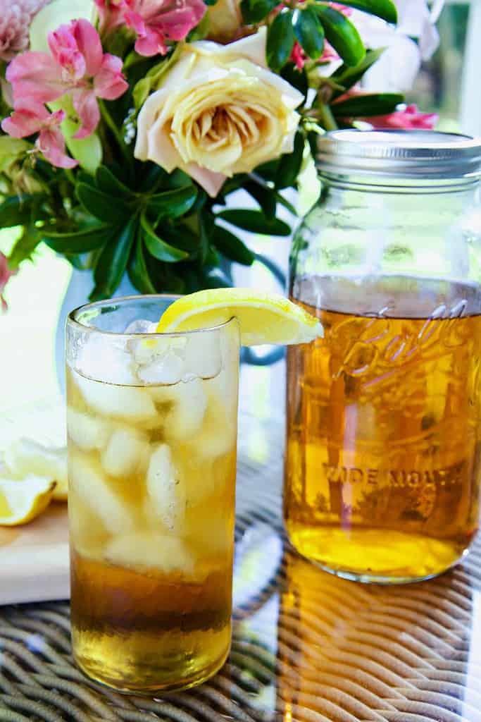 a tall glass of sun tea with ice and a lemon wedge flowers and large mason jar with tea in background