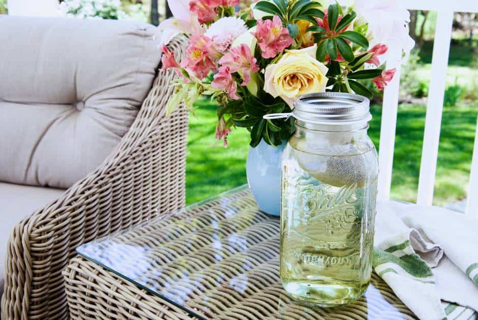 large mason jar steeping tea on a wicker table outside flowers and chair in background