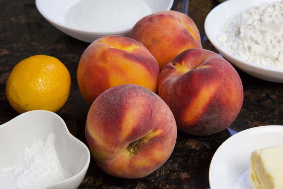 four fresh ripe peaches and a lemon surrounded by ingredients to make a peach cobbler