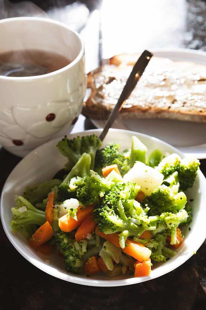 small white bowl with steamed broccoli carrots with buttered toast and a cup of tea