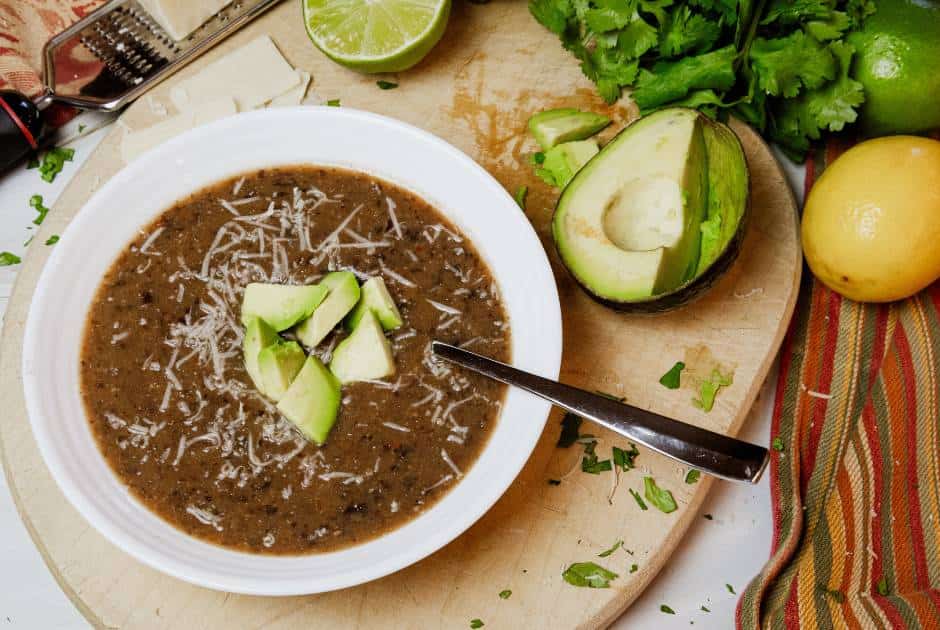 bowl of black bean soup with sliced avocado and cilantro on a cutting board