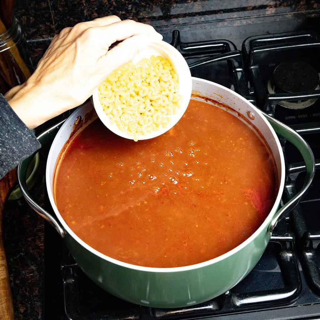 large soup pot on the stove with chili and a woman hand holding a small white bowl of pasta getting ready to pour it in the pot