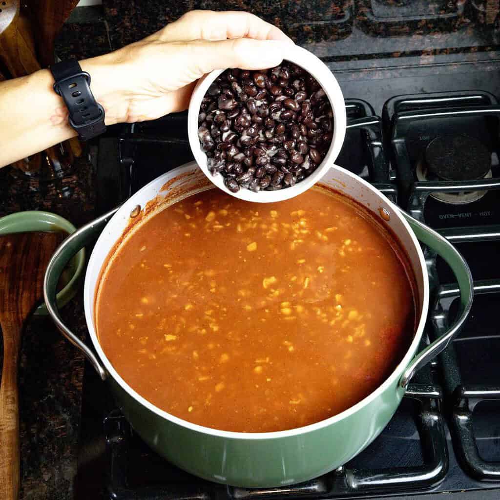 a womans hand holding a small white bowl with black beans getting ready to pour it in the large soup pot on the stove