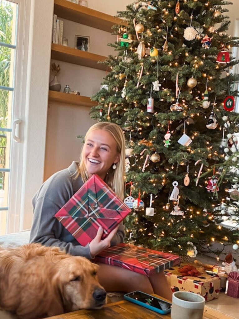 young woman sitting in front of a christmas tree holding a present laughing with her dog lying by her side